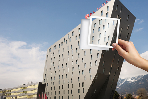 Ein graues Hotelgebäude mit blauem Himmel. Eine Hand hält ein durchsichtiges Quadrat davor und fokussiert das Gebäude.
