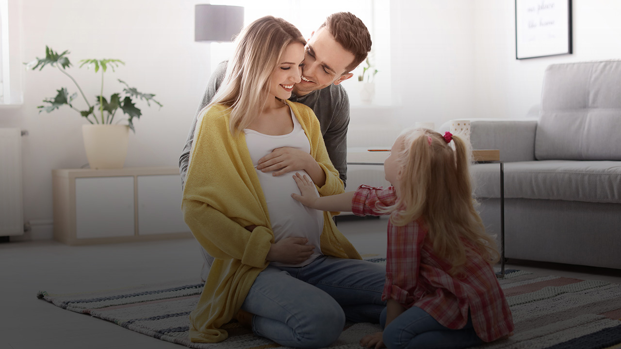 Eine schwangere Frau sitzt mit ihrem Mann und ihrer Tochter auf dem Boden. Das Mädchen berührt den Babybauch.