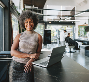 Eine Frau mit Afro im Business-Dress lehnt sich an einen Tisch und lächelt. Auf dem Tisch befindet sich ein Laptop.