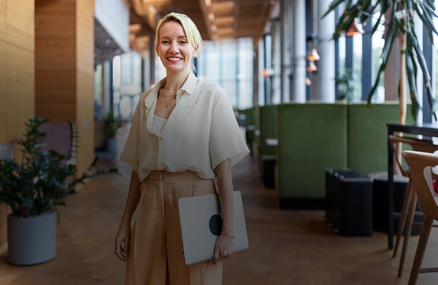 Eine Frau mit blondem kurzen Haar steht in einem Büro mit Laptop unter dem Arm.
