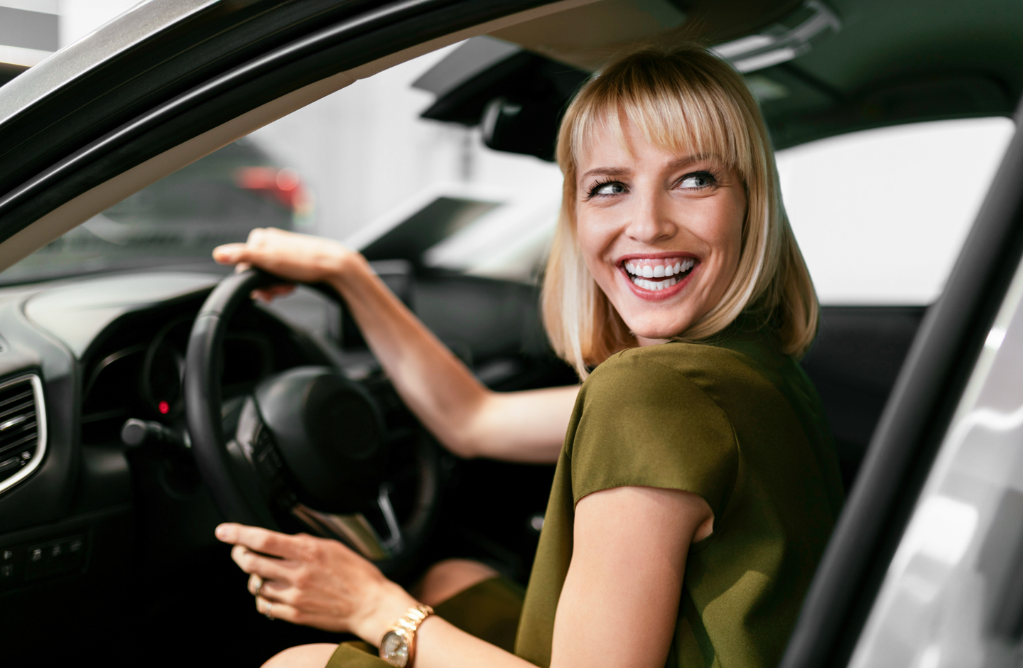 Eine Dame sitzt hinter dem Steuer ihres Autos und lacht freundlich.